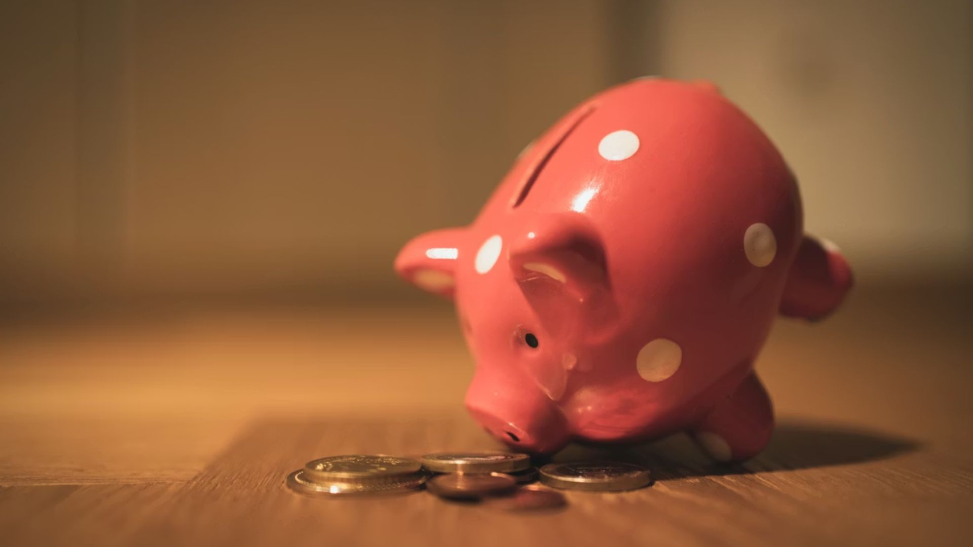 Pink polka-dotted piggy bank leaning forward toward a small scattering of coins on a wooden surface.
