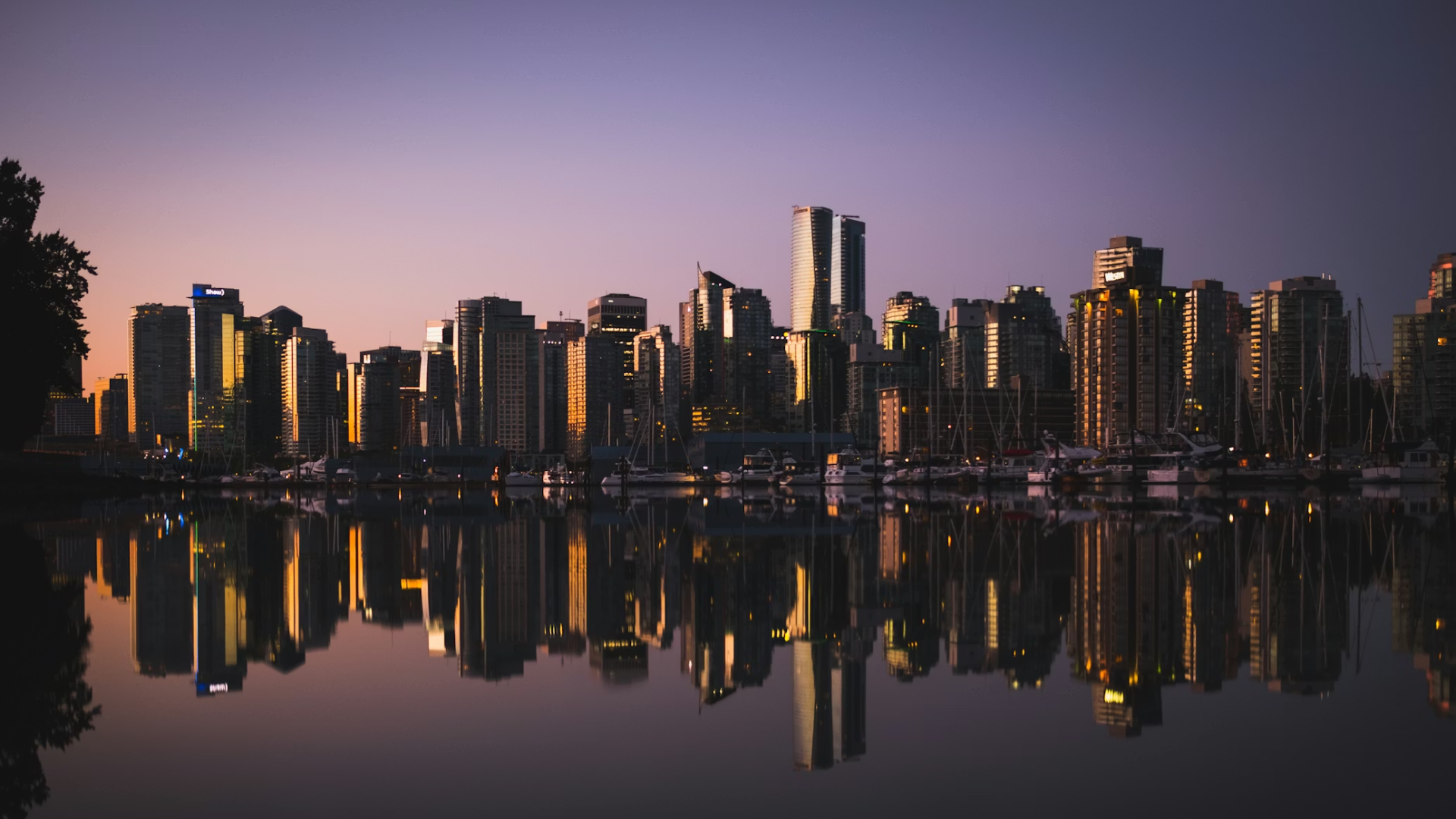 A city skyline at dusk with the tall buildings reflected perfectly in the still water of a harbor in the foreground.