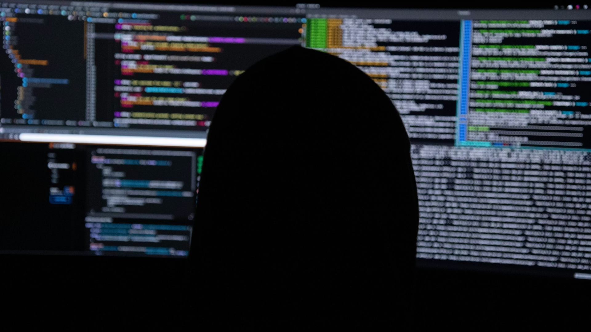 The silhouette of a person wearing a hoodie, seen from behind, looking at multiple computer monitors filled with lines of code.