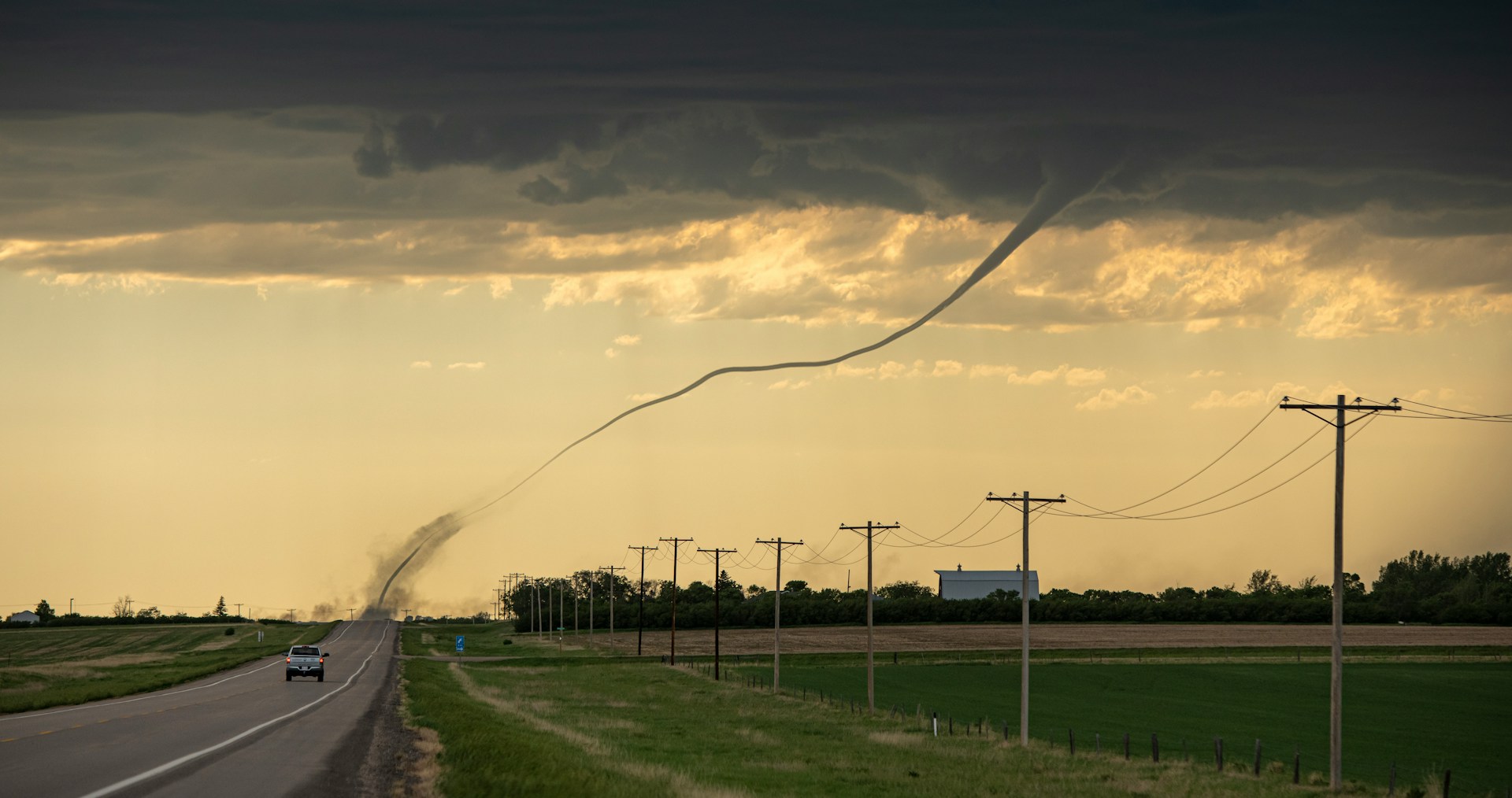 Large tornado funnel stretching across the sky over farmland near a rural road.