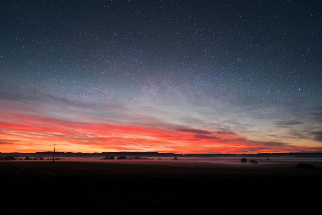 Star-filled night sky above a red and orange sunset over a rural landscape.