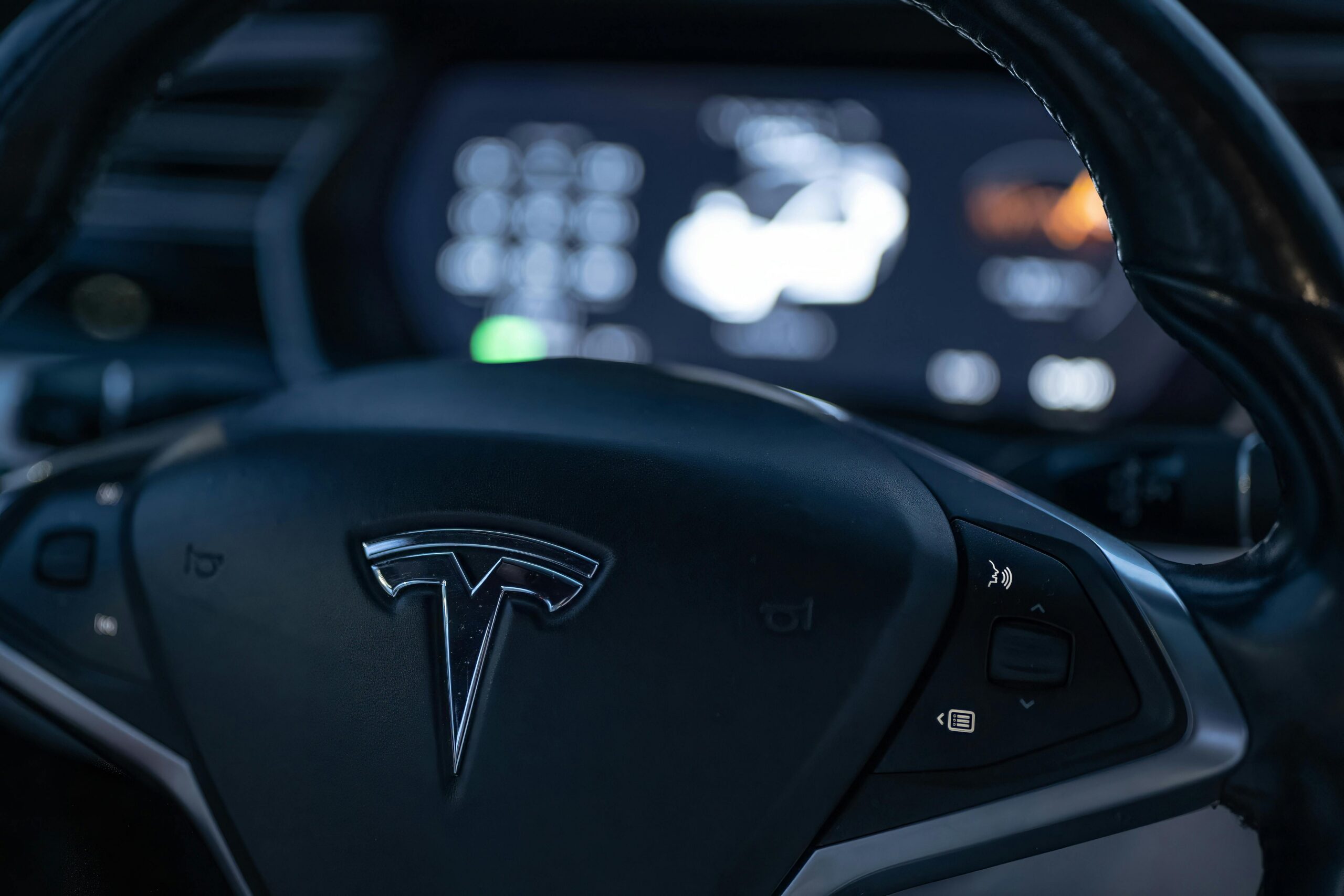 Tesla steering wheel with illuminated Tesla logo and digital dashboard display in the background.
