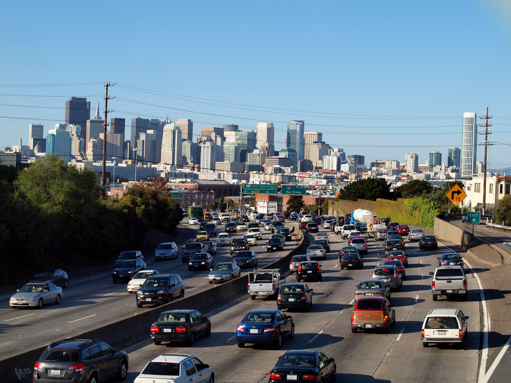 Heavy traffic on a multi-lane freeway with the San Francisco skyline visible in the background.