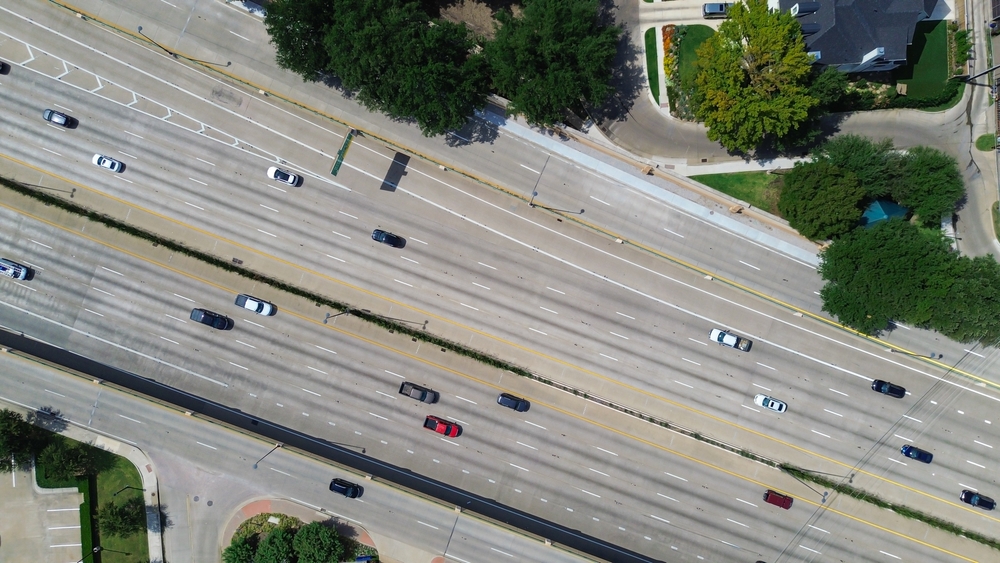 Aerial view of a multi-lane highway with dense traffic passing residential homes and trees nearby.