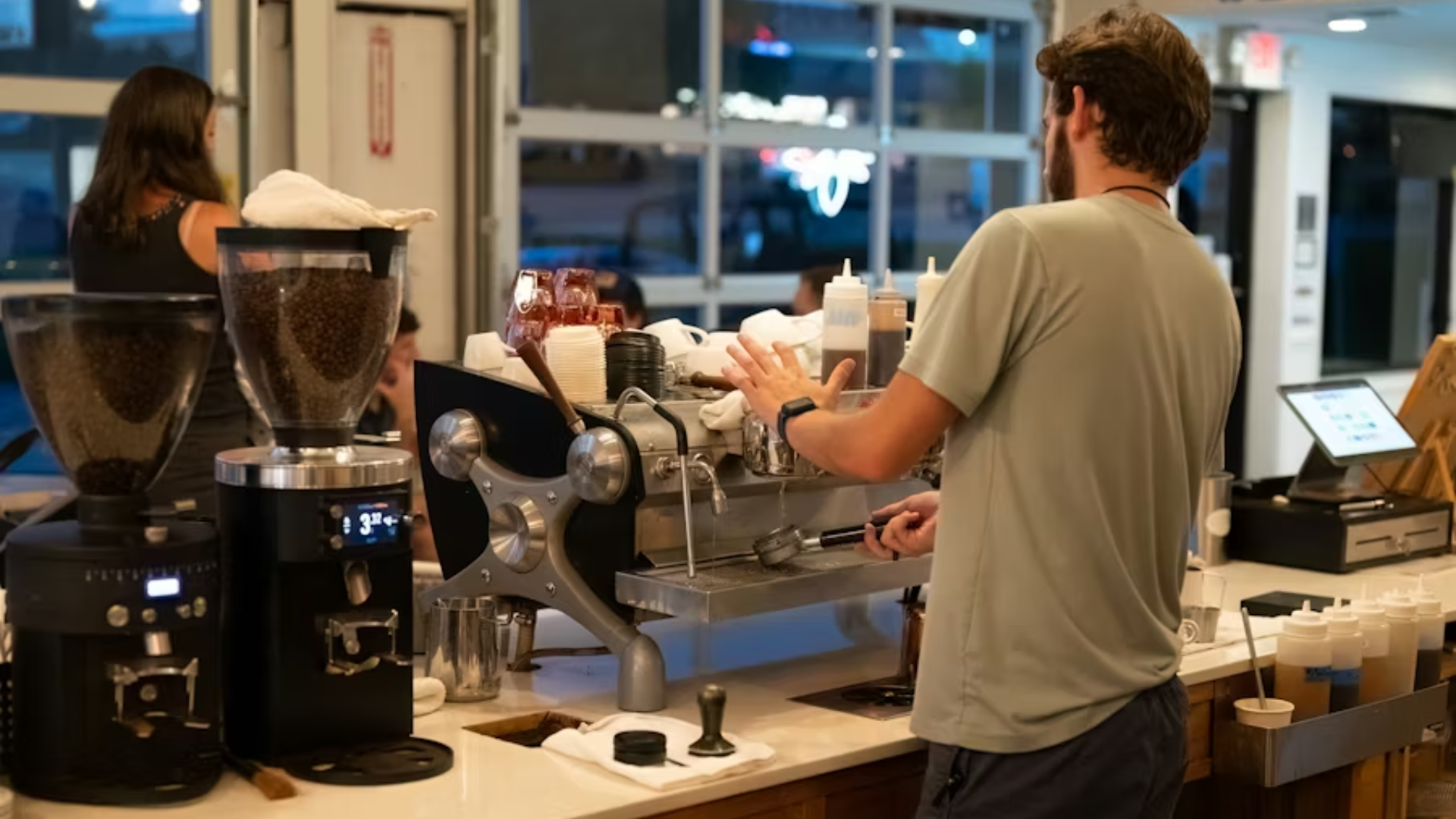 A medium shot from behind a bearded barista in a grey t-shirt as he operates a high-end chrome espresso machine.