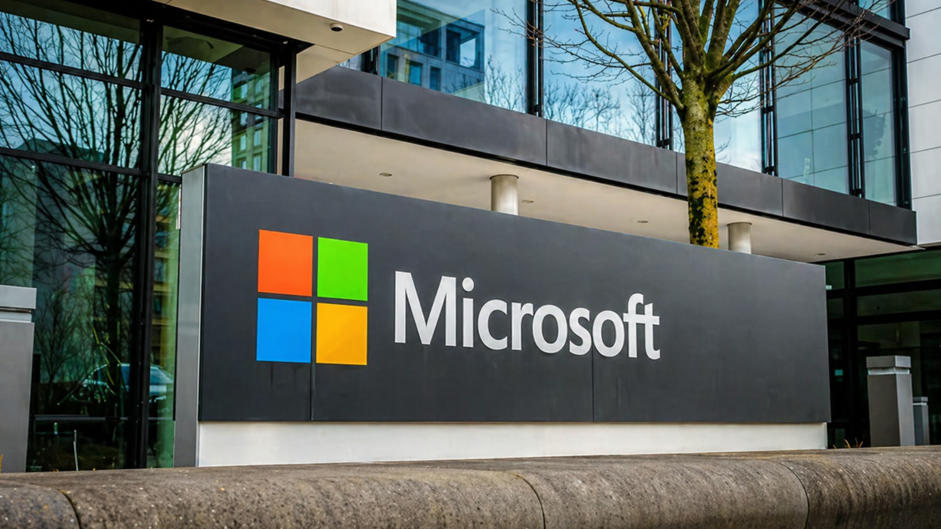 A daytime shot of the Microsoft corporate signage featuring the classic four-color window logo (orange, green, blue, yellow) in front of a modern glass-and-metal headquarters building; a bare tree and building reflections are visible in the windows.