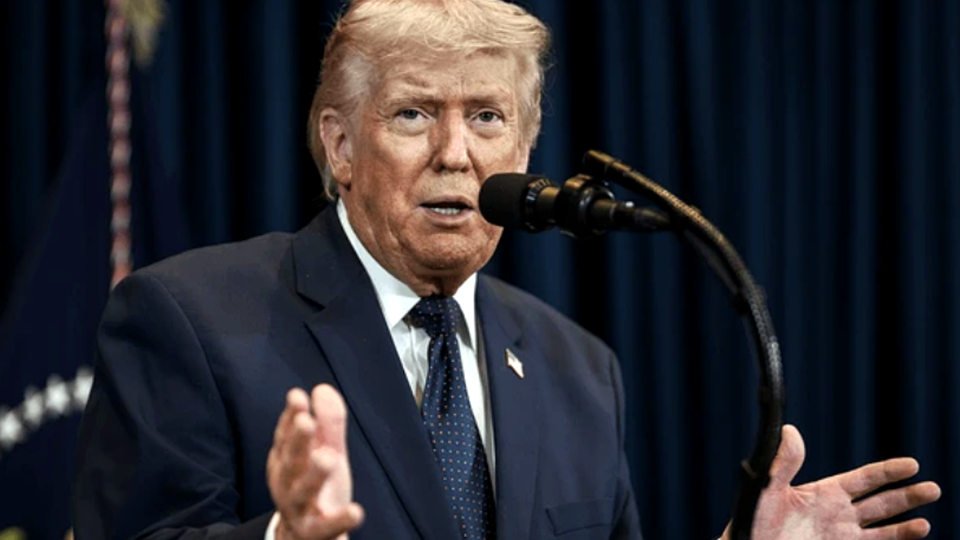 A close-up of Donald Trump speaking into a microphone with his hands raised in an expressive gesture; he is wearing a navy blue suit and a polka-dot tie against a backdrop of deep blue curtains and a partially visible American flag.