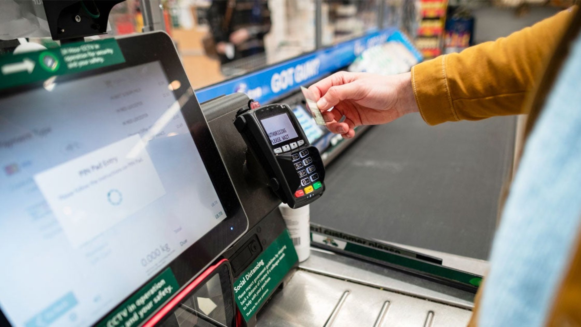 A high-angle view of a self-checkout station in a grocery store, showing a customer's hand inserting a credit card into a payment terminal.