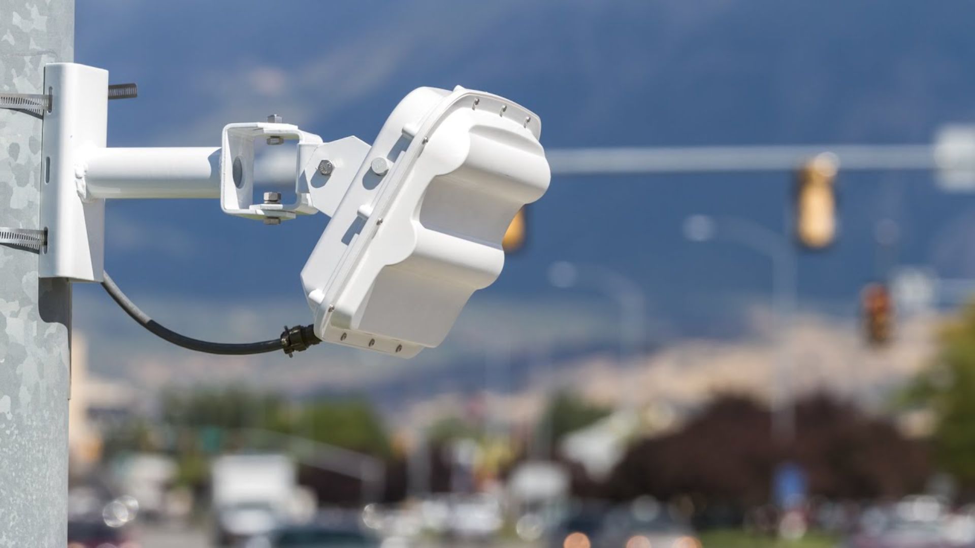 Side view of a white, wavy traffic-detection sensor mounted on a roadside pole, with blurred cars and traffic lights in the background.