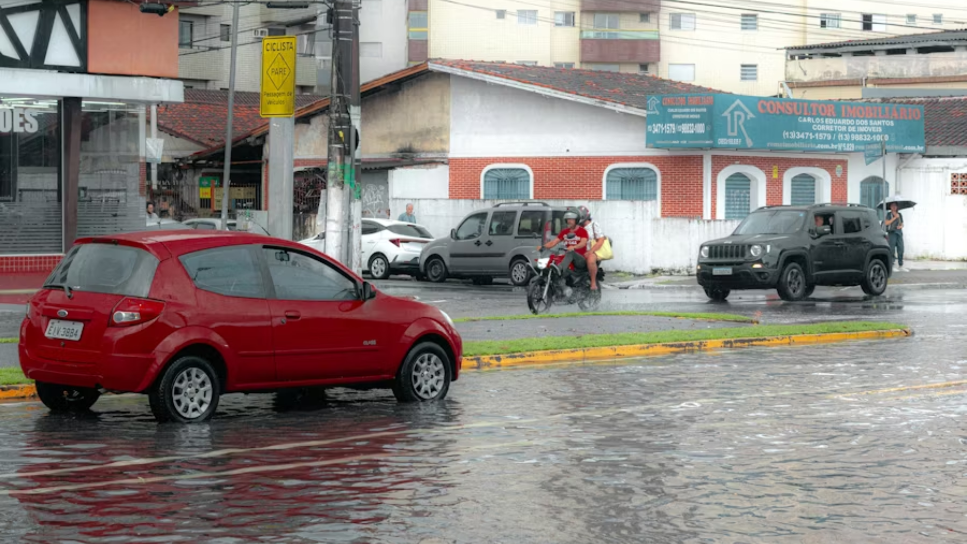 A red Ford Ka is parked on a flooded city street in a residential area, with water reaching halfway up its tires; a motorcyclist and an SUV navigate the deep puddles under a grey, rainy sky.
