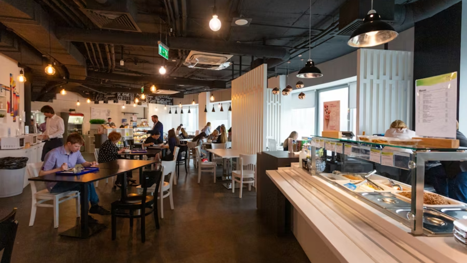 A wide interior shot of a brightly lit, modern restaurant with light wood tables, white chairs, and several people seated and eating.