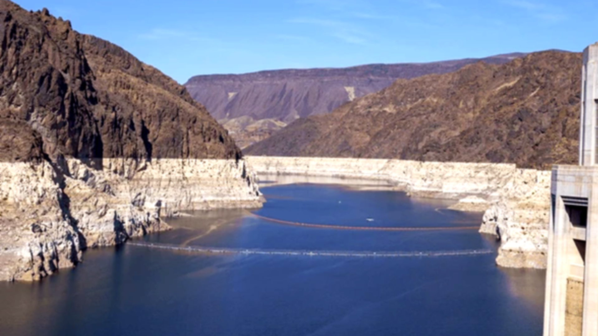 A high-angle shot of Lake Mead behind the Hoover Dam in April 2026; the water level is visibly low, revealing a massive, stark white "bathtub ring" of mineral deposits on the dark canyon walls, with floating safety barriers visible on the water's surface.