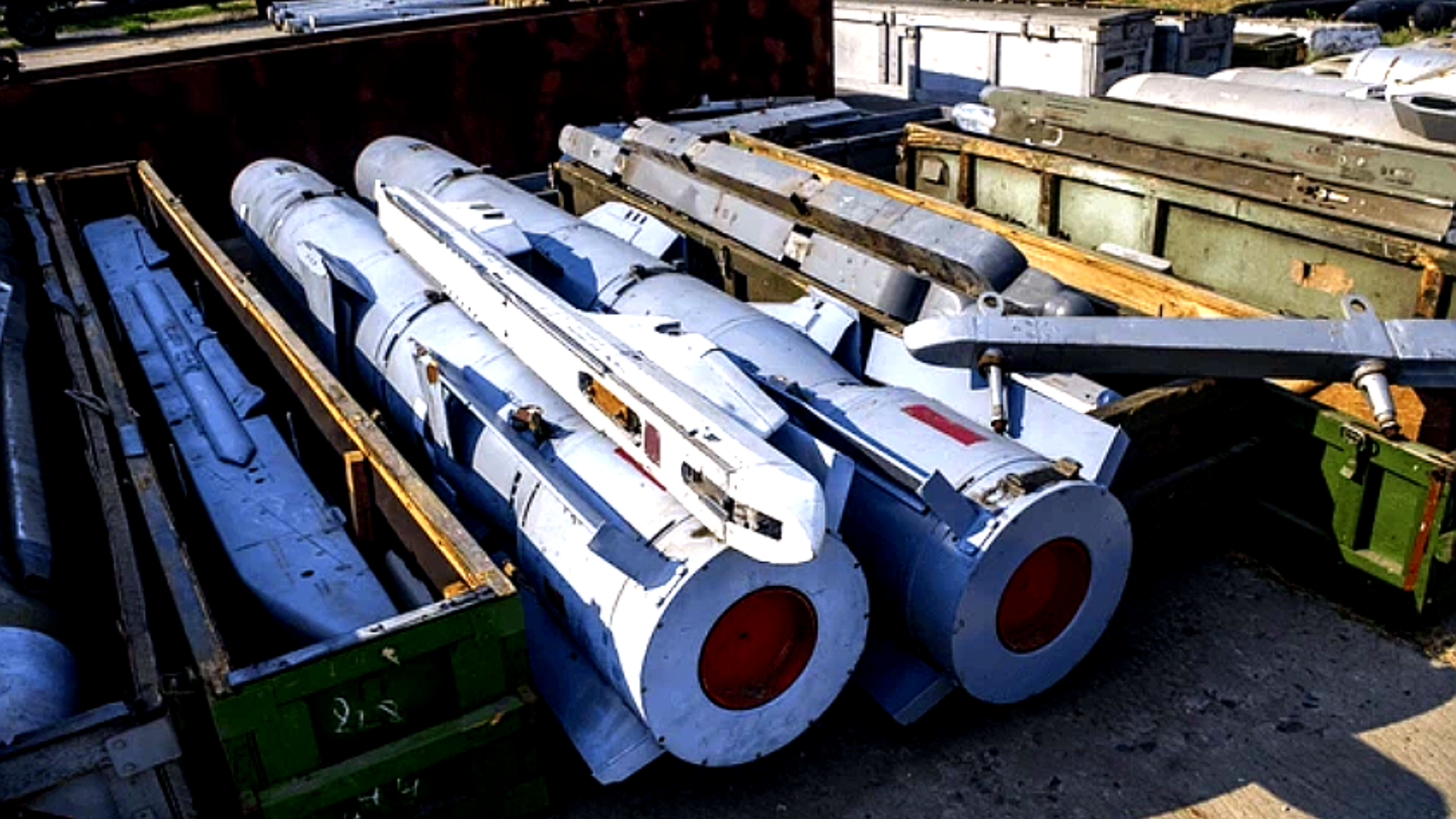 A high-angle shot of several large, cylindrical grey missiles resting in open green and metal storage crates at an outdoor military staging area, representing a stockpile of long-range munitions.
