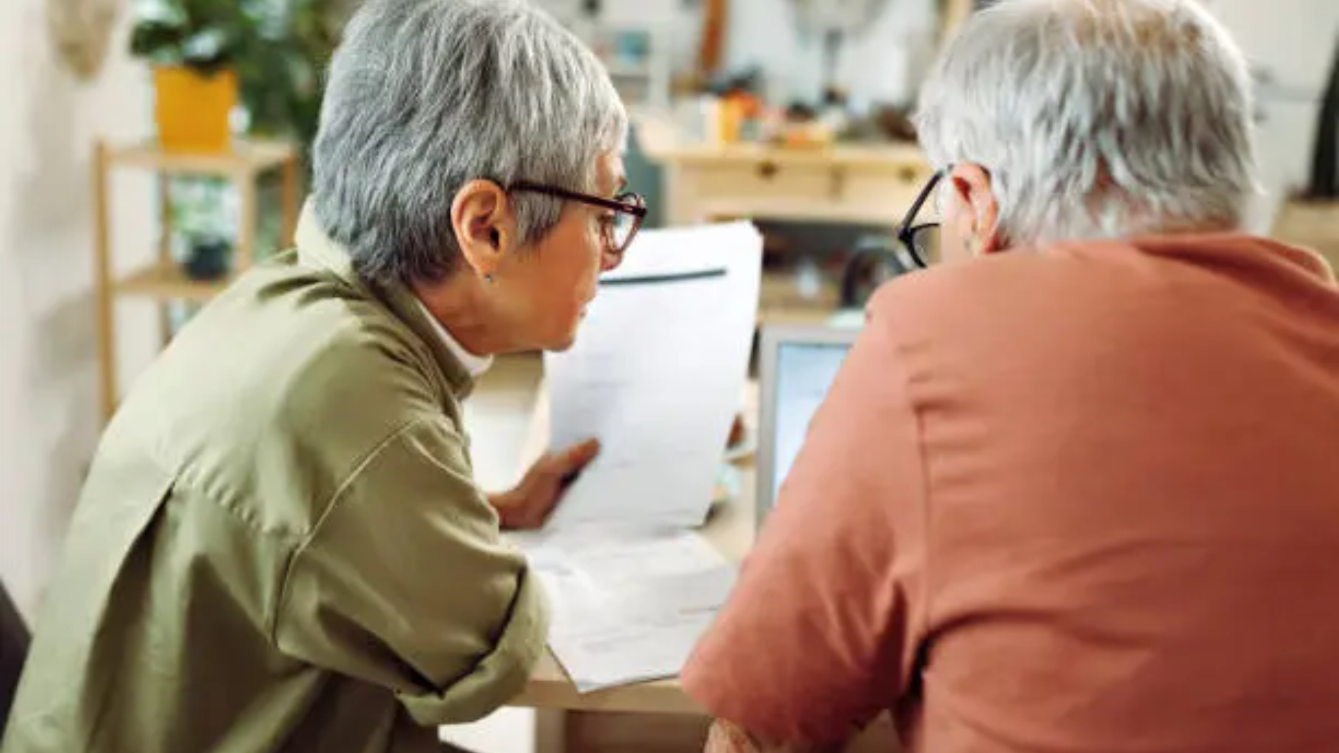 A high-angle shot from behind of an elderly couple with grey hair sitting at a wooden table; they are leaning in together to look at a laptop screen and several printed documents, symbolizing a consultation on personal finances or government benefits.