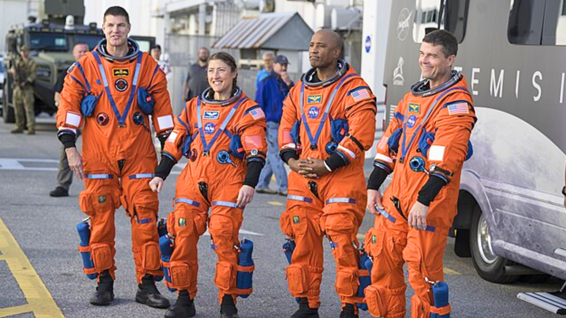 Four astronauts wearing bright orange spacesuits stand together outside near a vehicle.