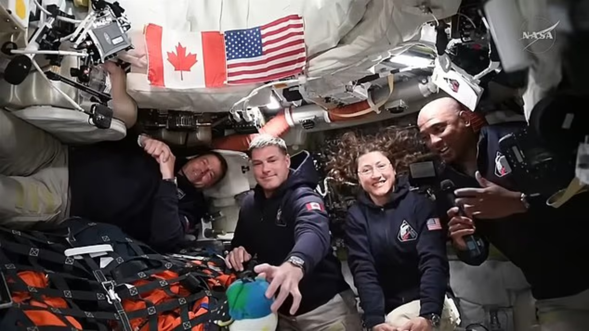 Four smiling astronauts float together inside a spacecraft under Canadian and American flags.