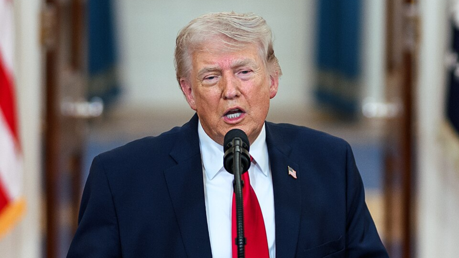 Donald Trump wearing a dark suit and red tie speaks into a microphone.