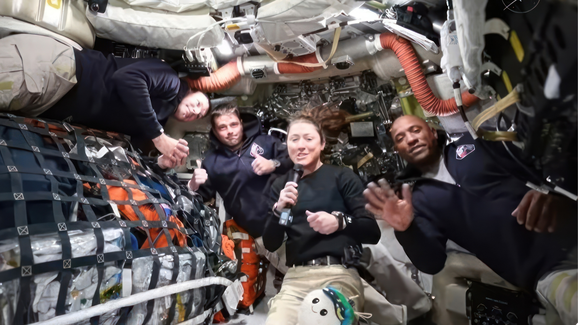 Four astronauts float inside a crowded spacecraft while one woman holds a microphone.
