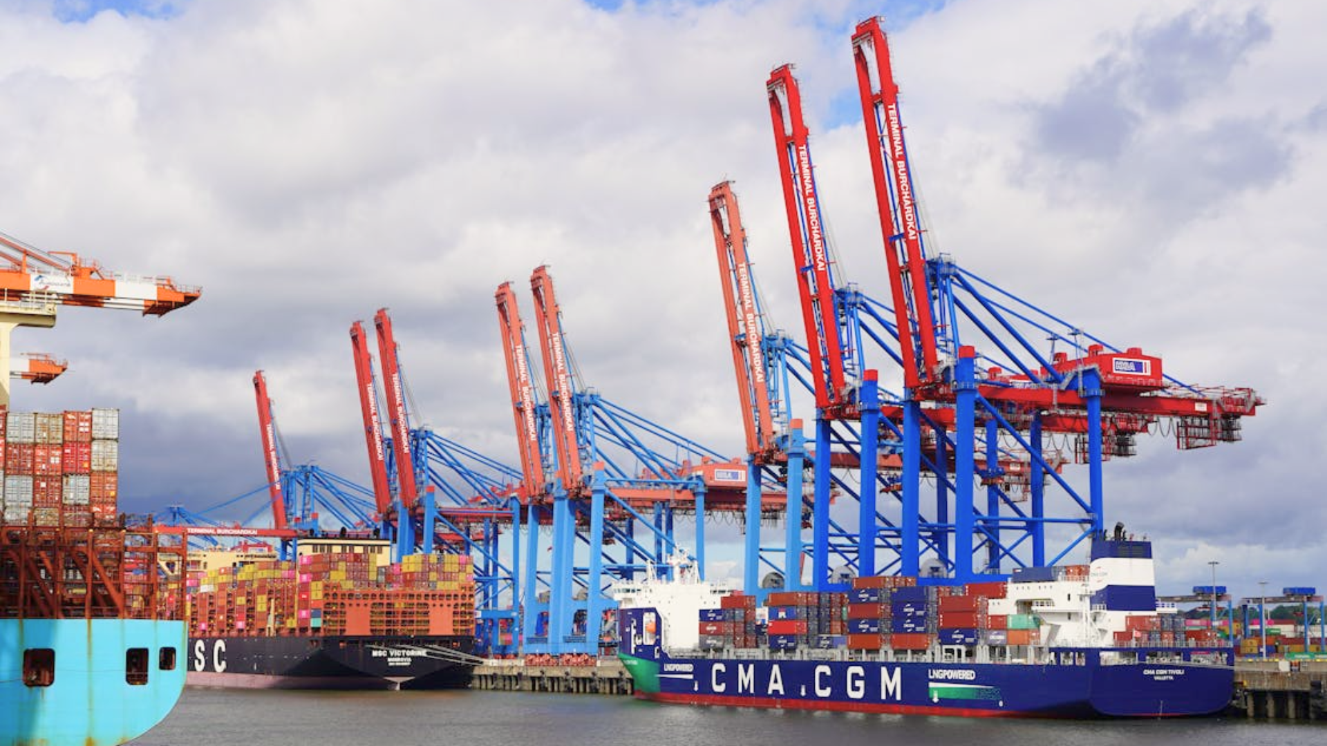 A large cargo ship loaded with colorful containers docked at a port with tall blue and red cranes.