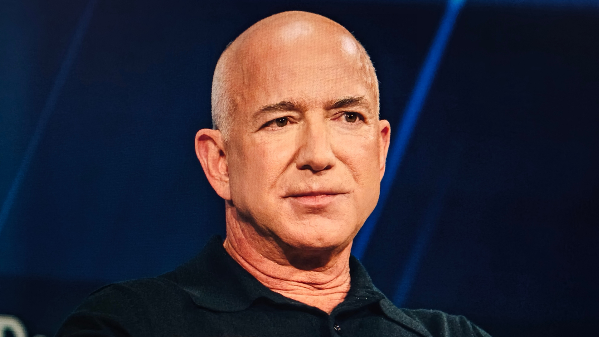 A close-up portrait of Jeff Bezos wearing a dark shirt against a blue background.