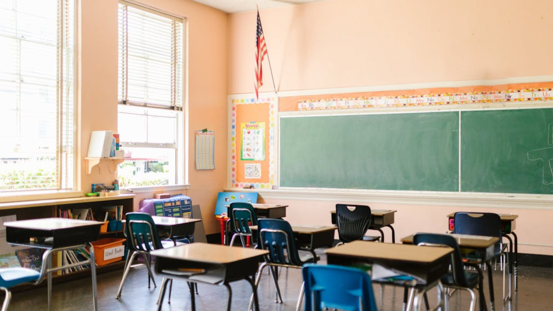An empty classroom with student desks, a green chalkboard, and an American flag.