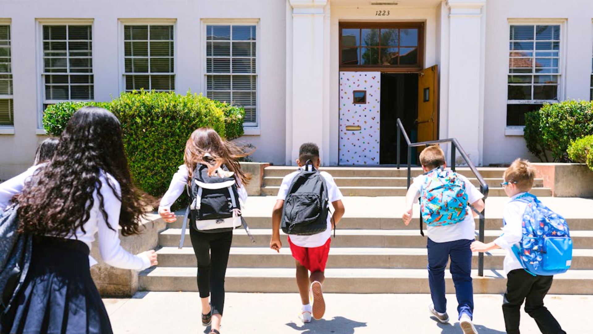 A group of kids with backpacks run up the steps to enter a school building.