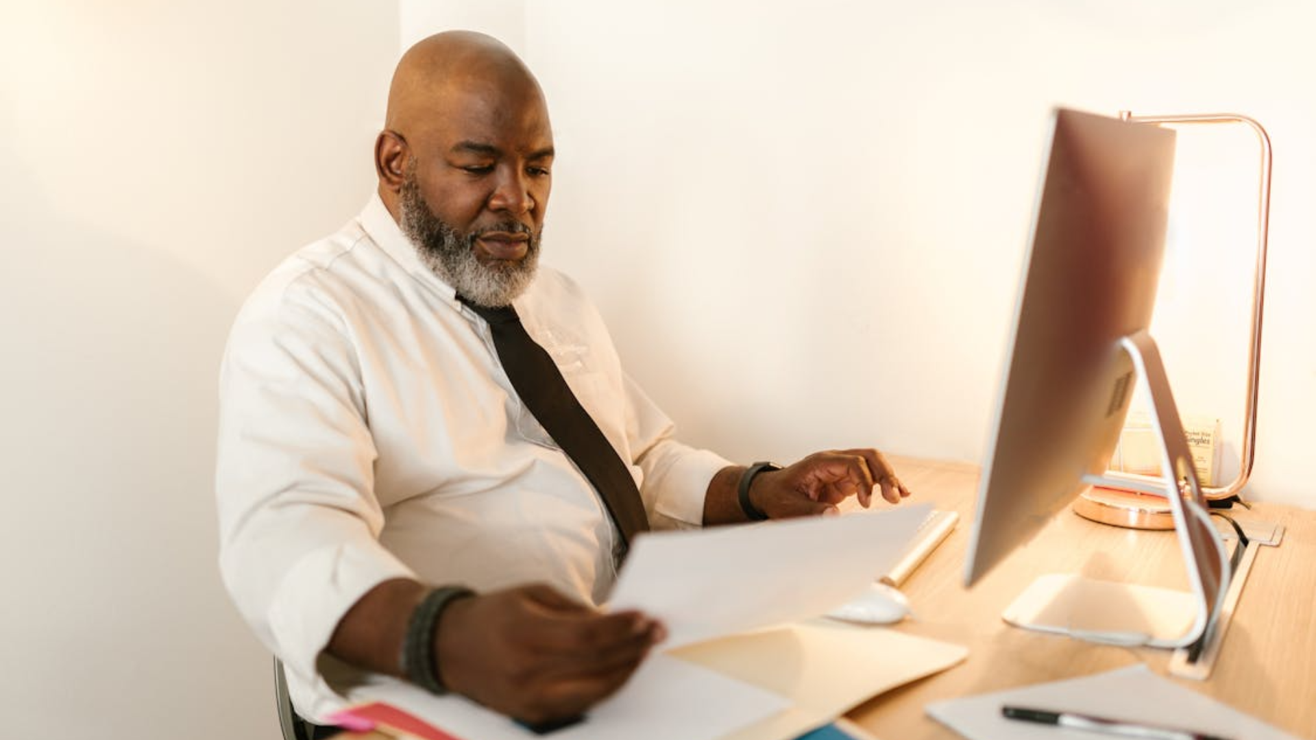 A man wearing a white shirt and tie sits at a desk looking at a document next to a computer.