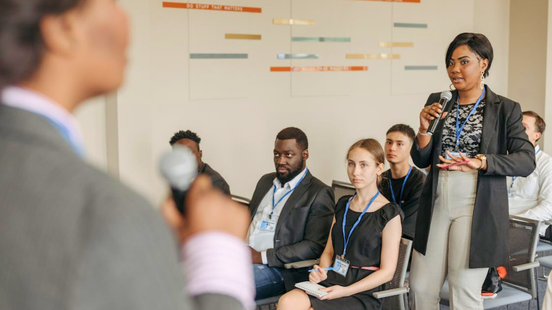 A woman stands with a microphone to speak to a seated audience at a meeting.