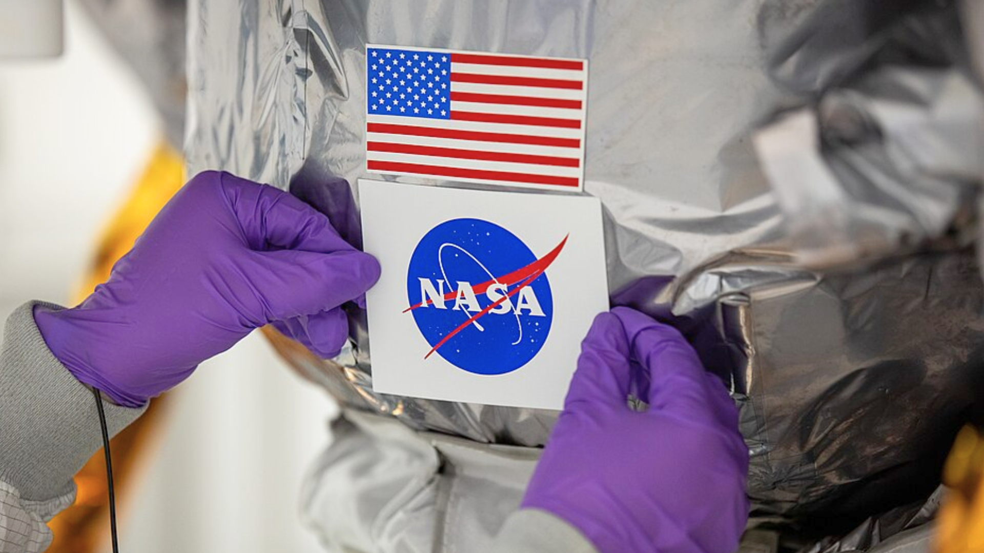 Hands wearing purple gloves place a NASA logo sticker below an American flag on a shiny silver surface.