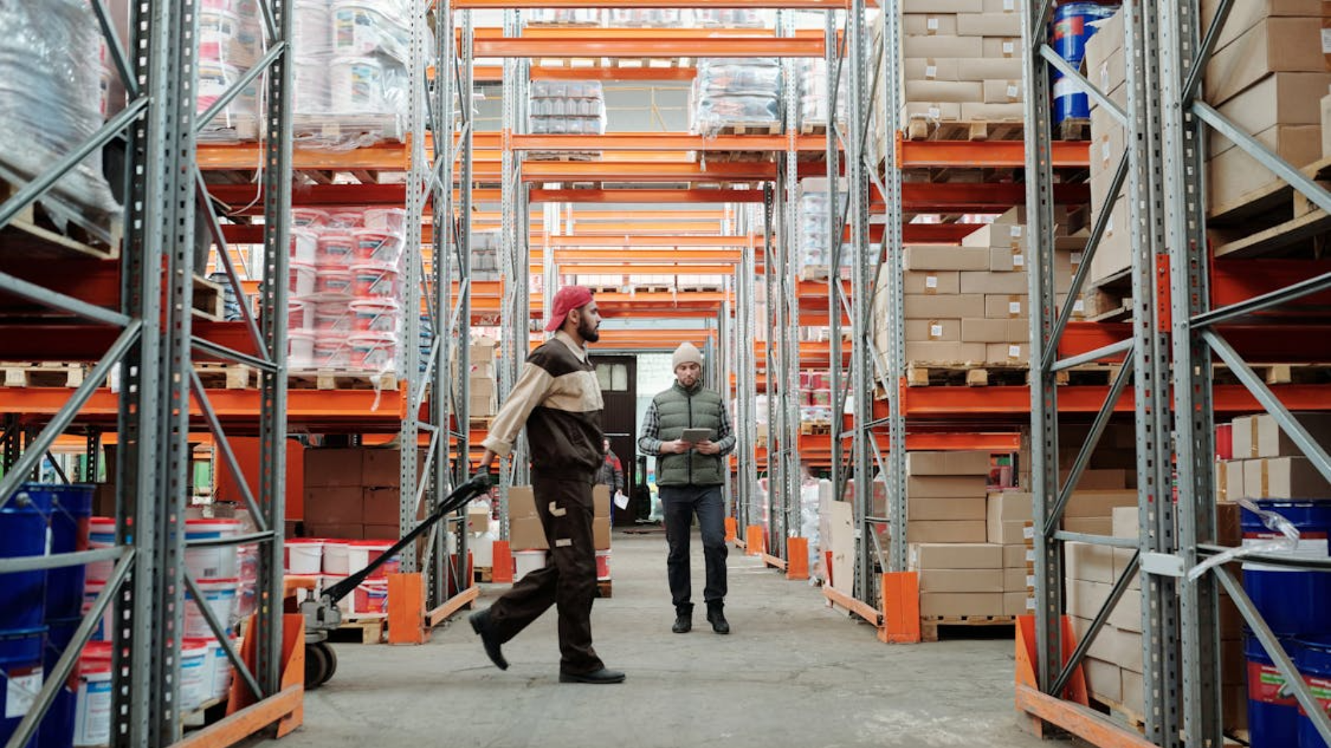 Two workers move through a large warehouse with tall orange shelves full of boxes and supplies.