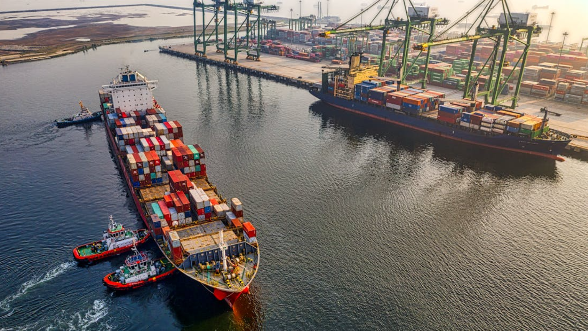 An aerial view of two tugboats guiding a large cargo ship filled with containers at a busy port.