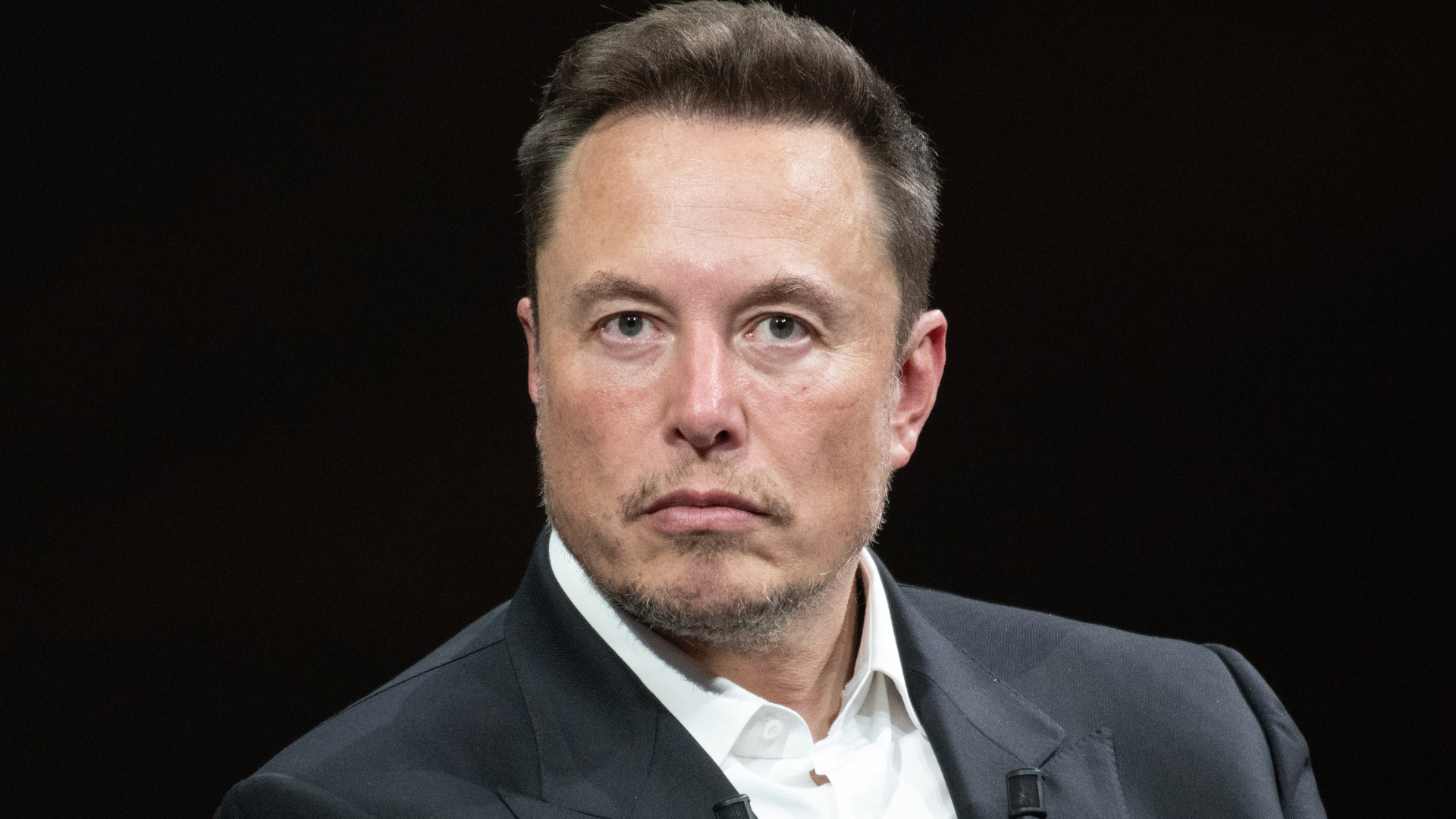 A close-up portrait of Elon Musk wearing a dark suit jacket and white shirt against a black background.