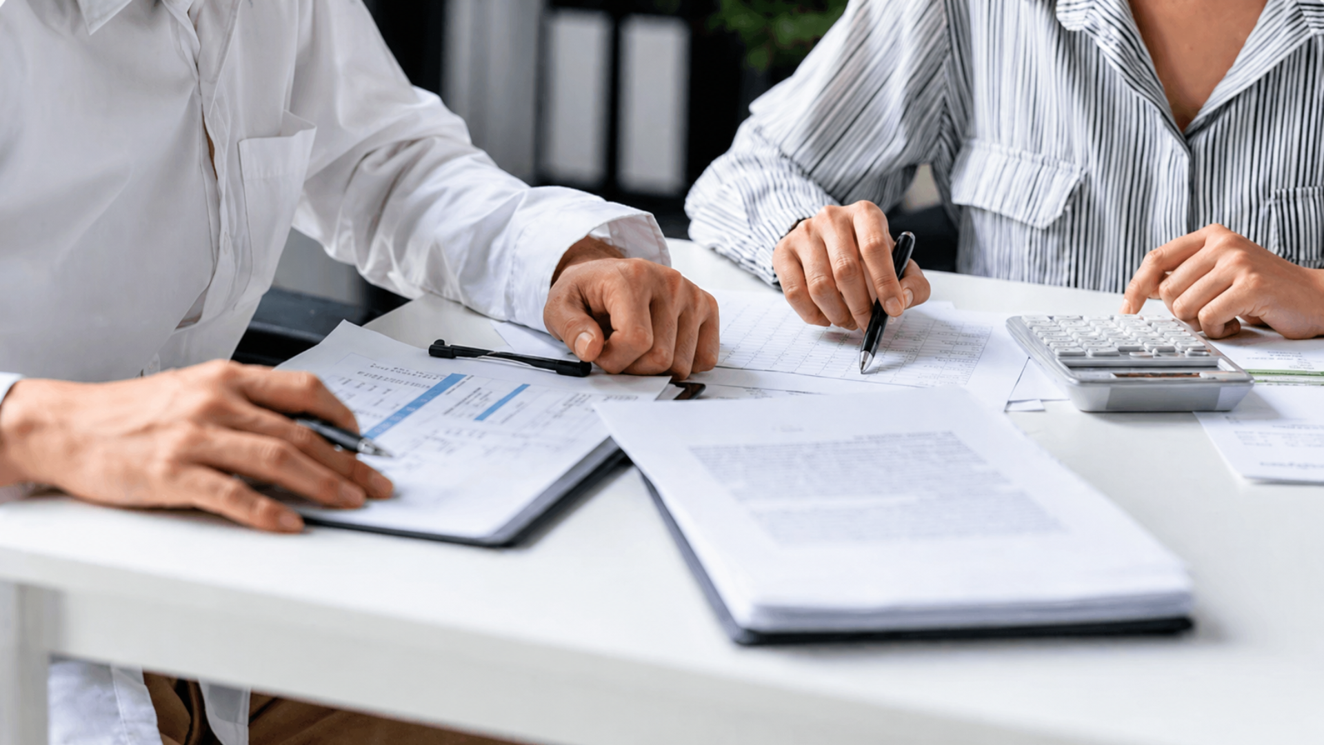 Two people review financial documents and use a calculator while discussing paperwork at a desk.