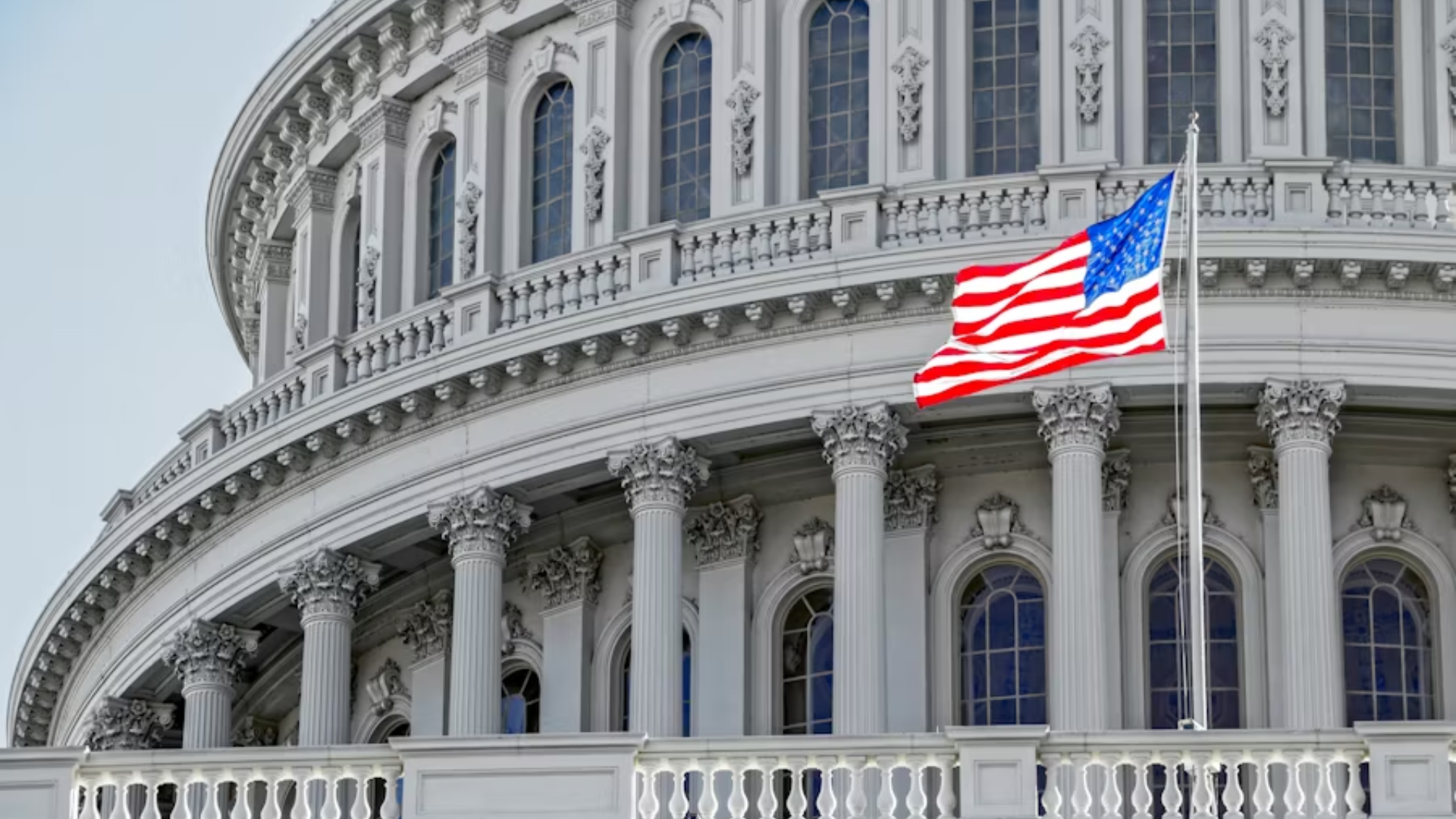 A close-up of the upper architectural details of the U.S. Capitol Building dome, featuring white columns and ornate carvings, with the American flag waving prominently in the foreground.