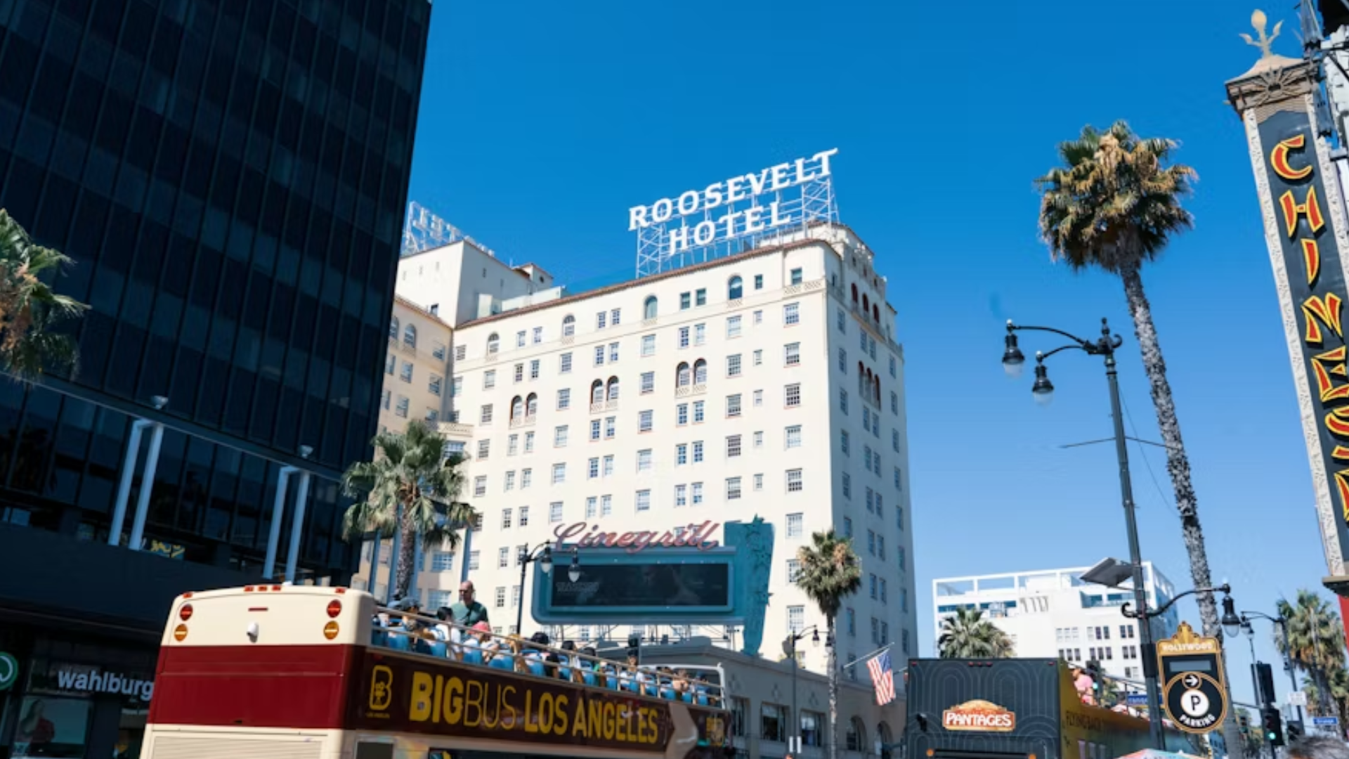 A daytime low-angle shot of the historic Roosevelt Hotel in Hollywood, Los Angeles, with its iconic white sign on the roof, palm trees in the foreground.