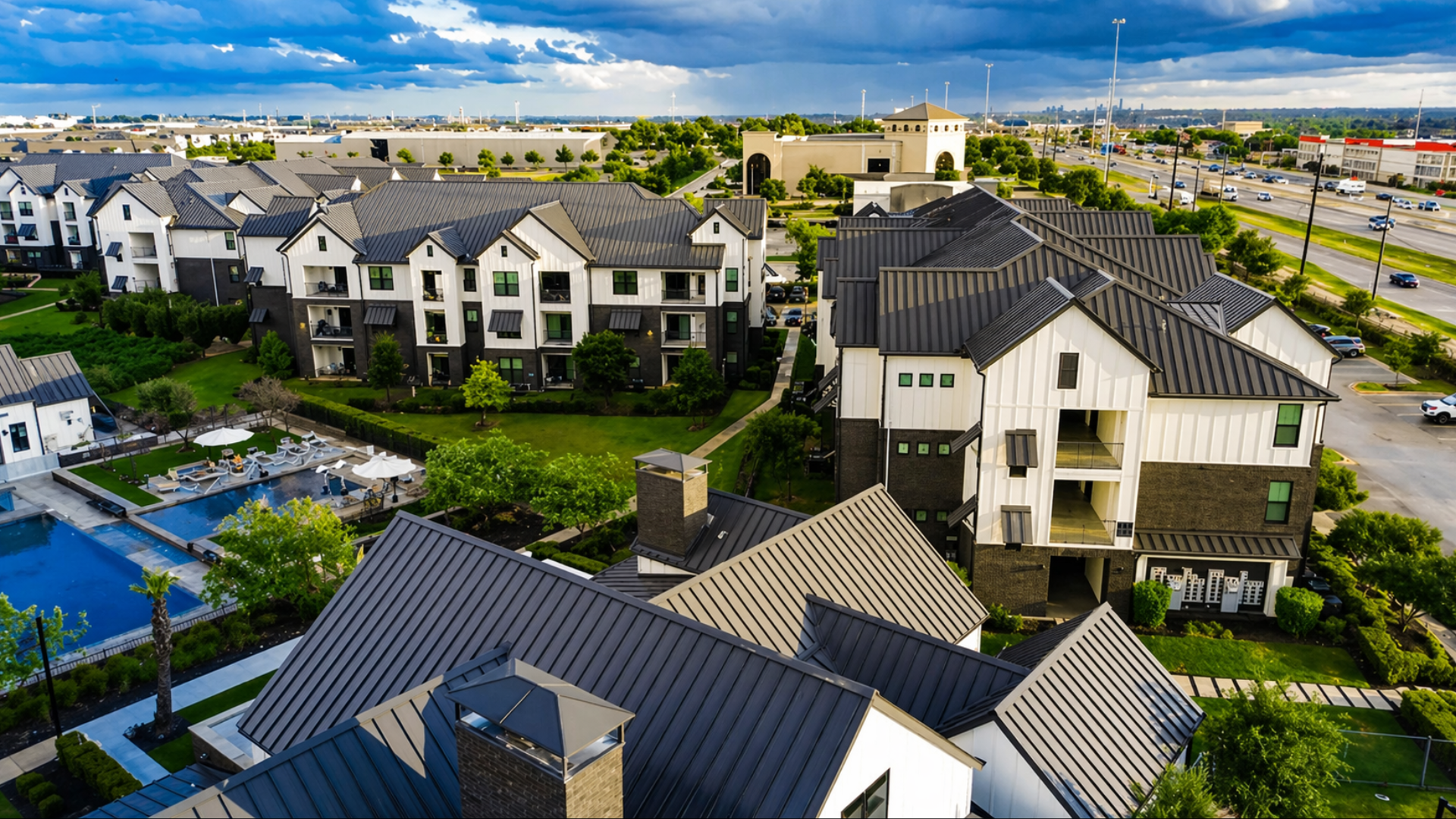 A high-angle view of a modern apartment complex with multiple buildings featuring white siding, black trim, and dark metal roofs; the complex includes a landscaped courtyard and a large blue swimming pool, situated next to a busy highway under a cloudy sky.