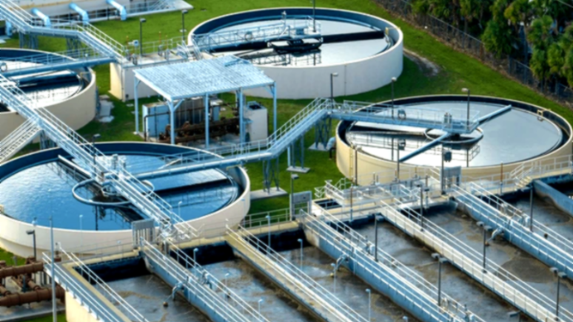 A top-down aerial view of a large wastewater treatment plant, featuring several circular blue clarification tanks connected by white walkways and industrial piping, set against a green manicured lawn.