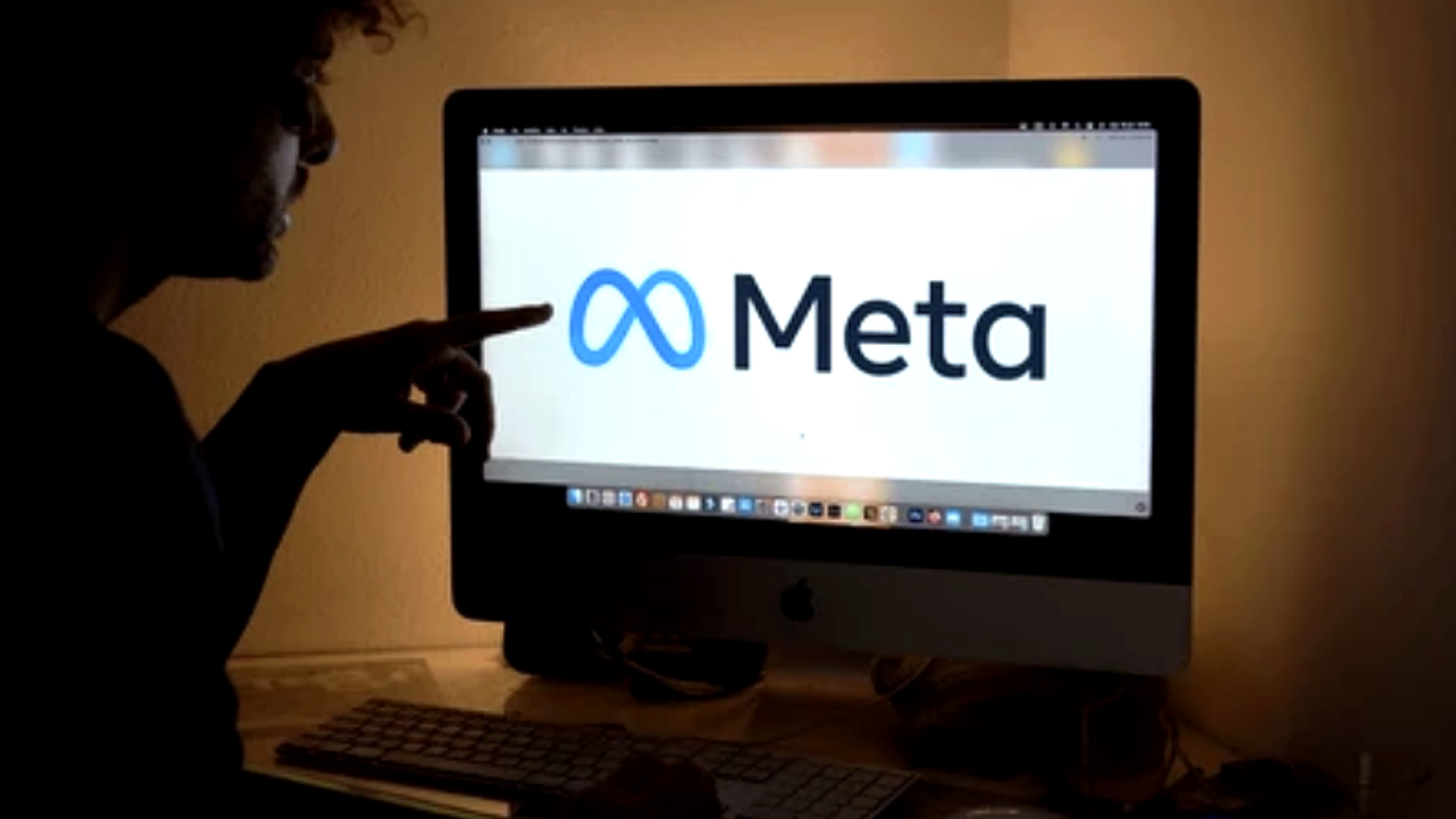 A shot in a dimly lit room where a person is pointing at a desktop monitor displaying the blue-and-white Meta logo; the screen is bright against the surrounding shadows, symbolizing the company's digital presence.