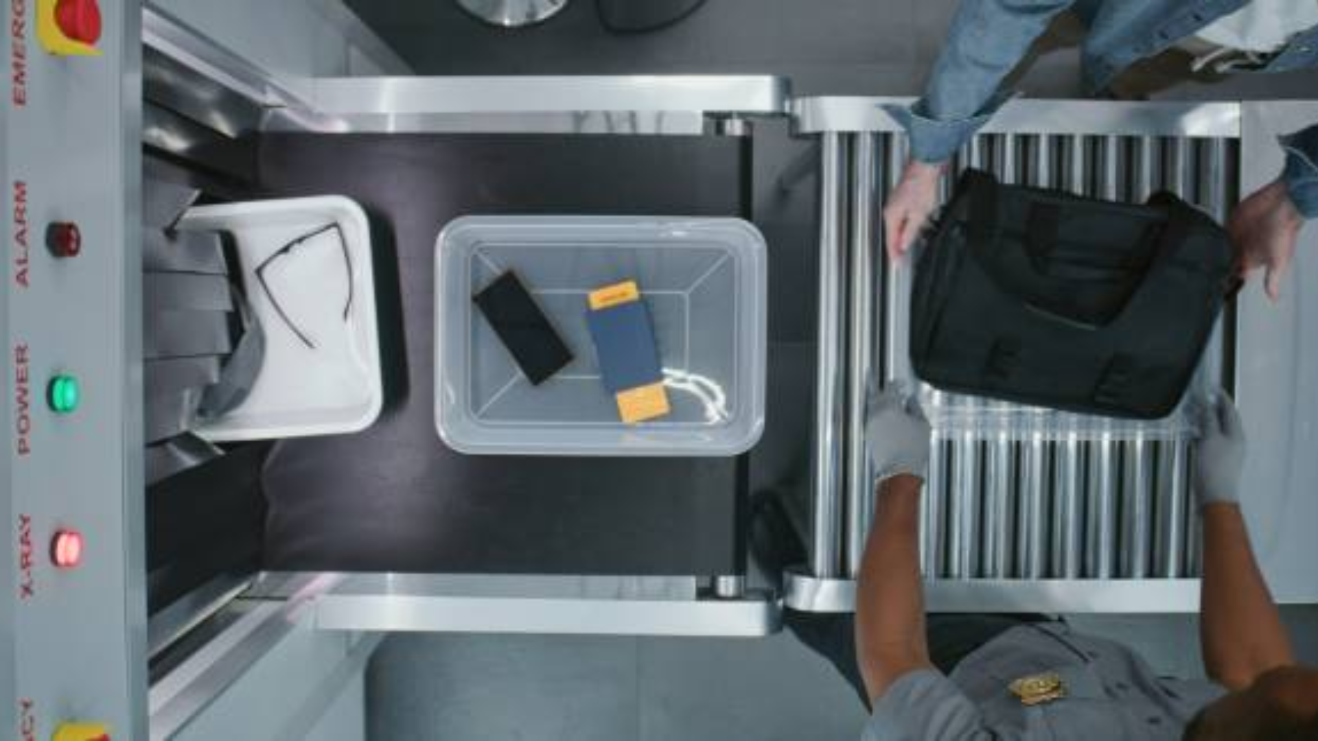 An overhead view of a security conveyor belt where plastic bins containing a passport, smartphone, and glasses are being moved toward an X-ray machine by a gloved agent.