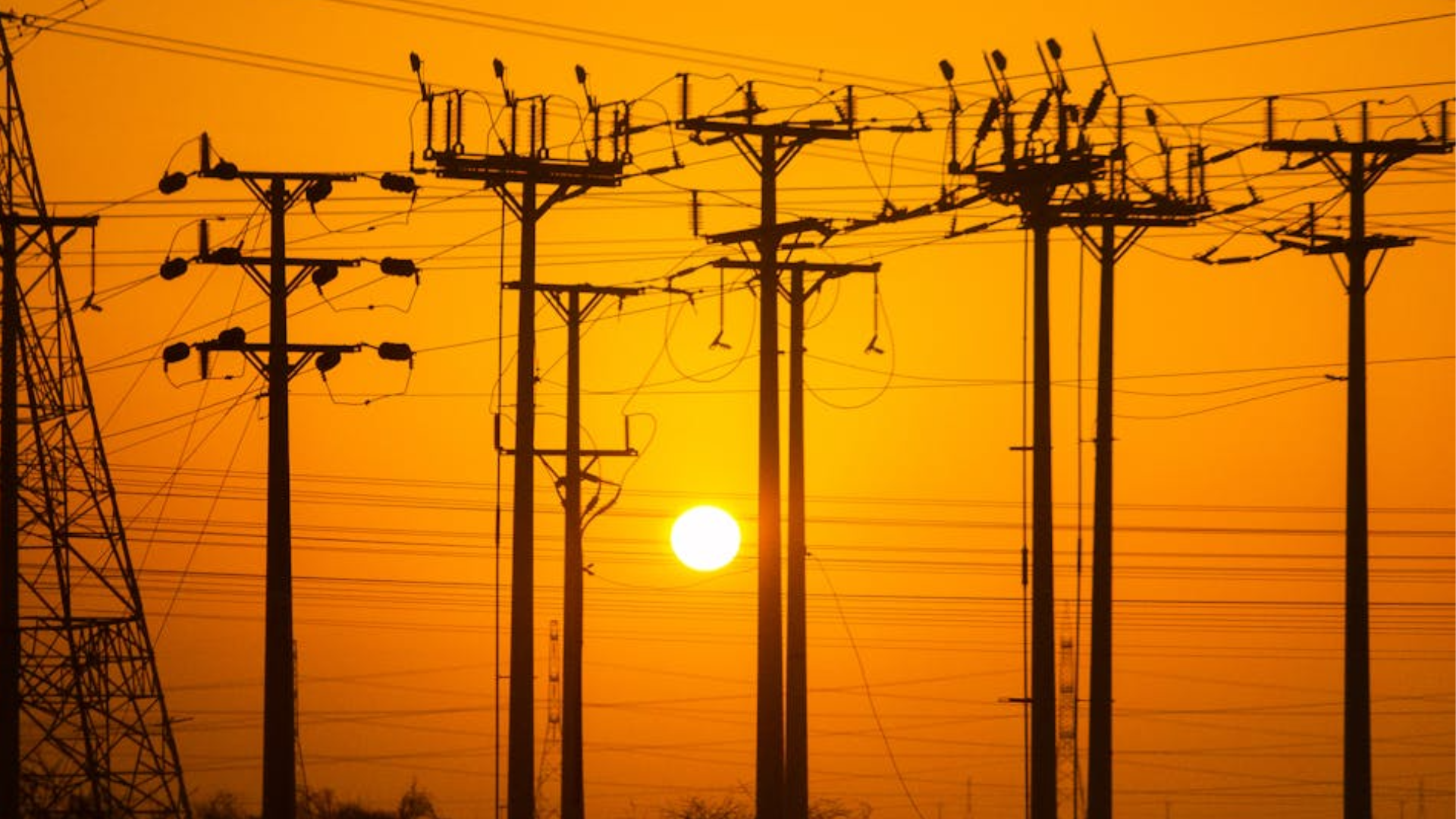 Dark silhouettes of electrical power lines and poles stand against a bright orange sunset.