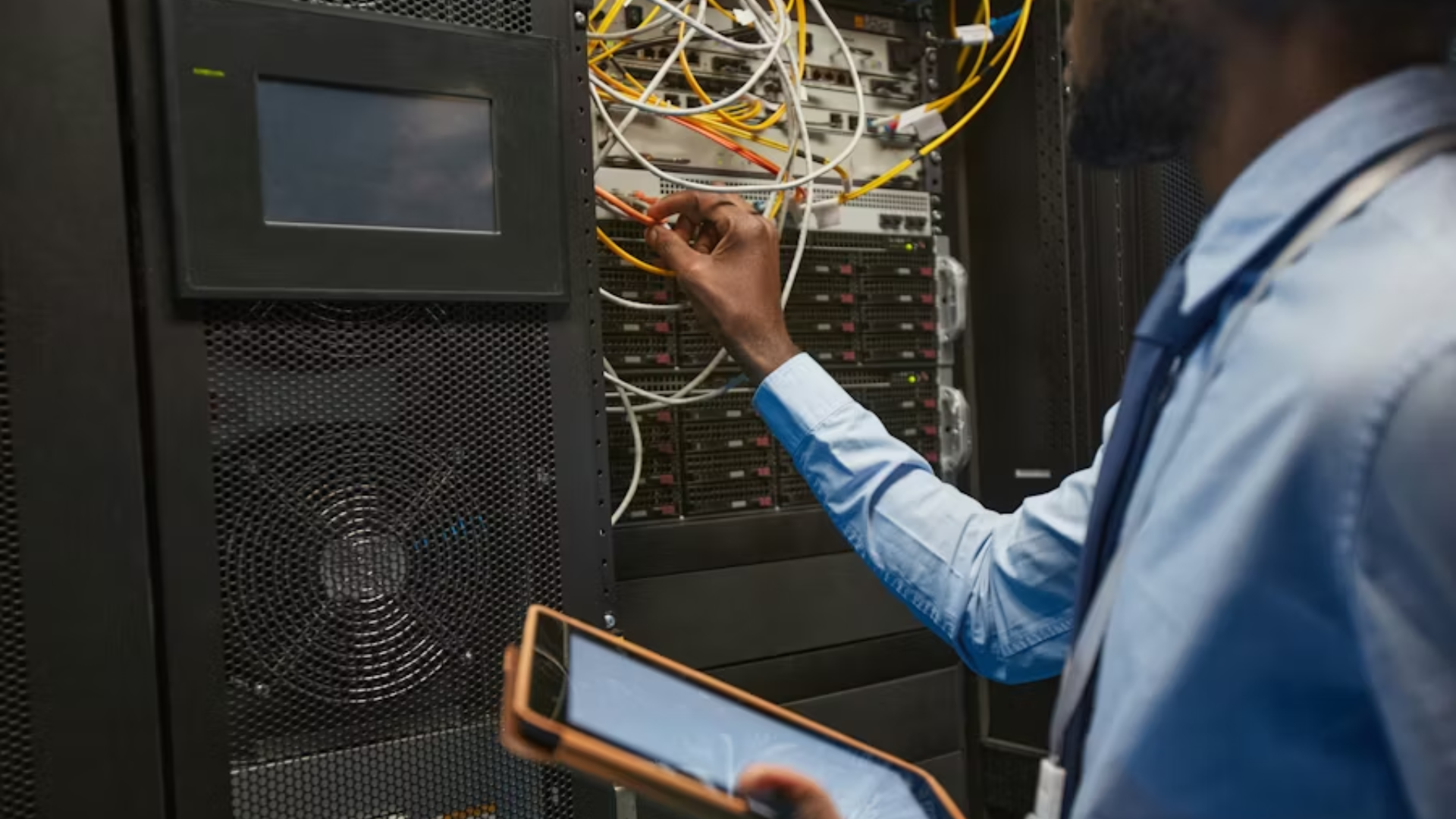 Technician connecting cables in a server rack while holding a tablet in a data center.