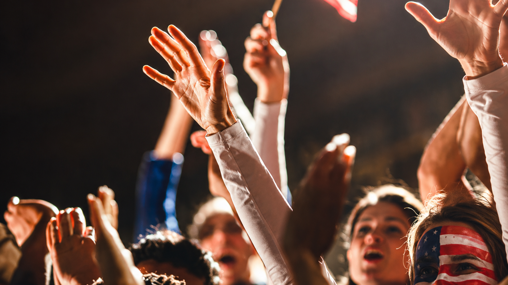 Crowd with raised hands and a person with American flag face paint cheering at a nighttime event.