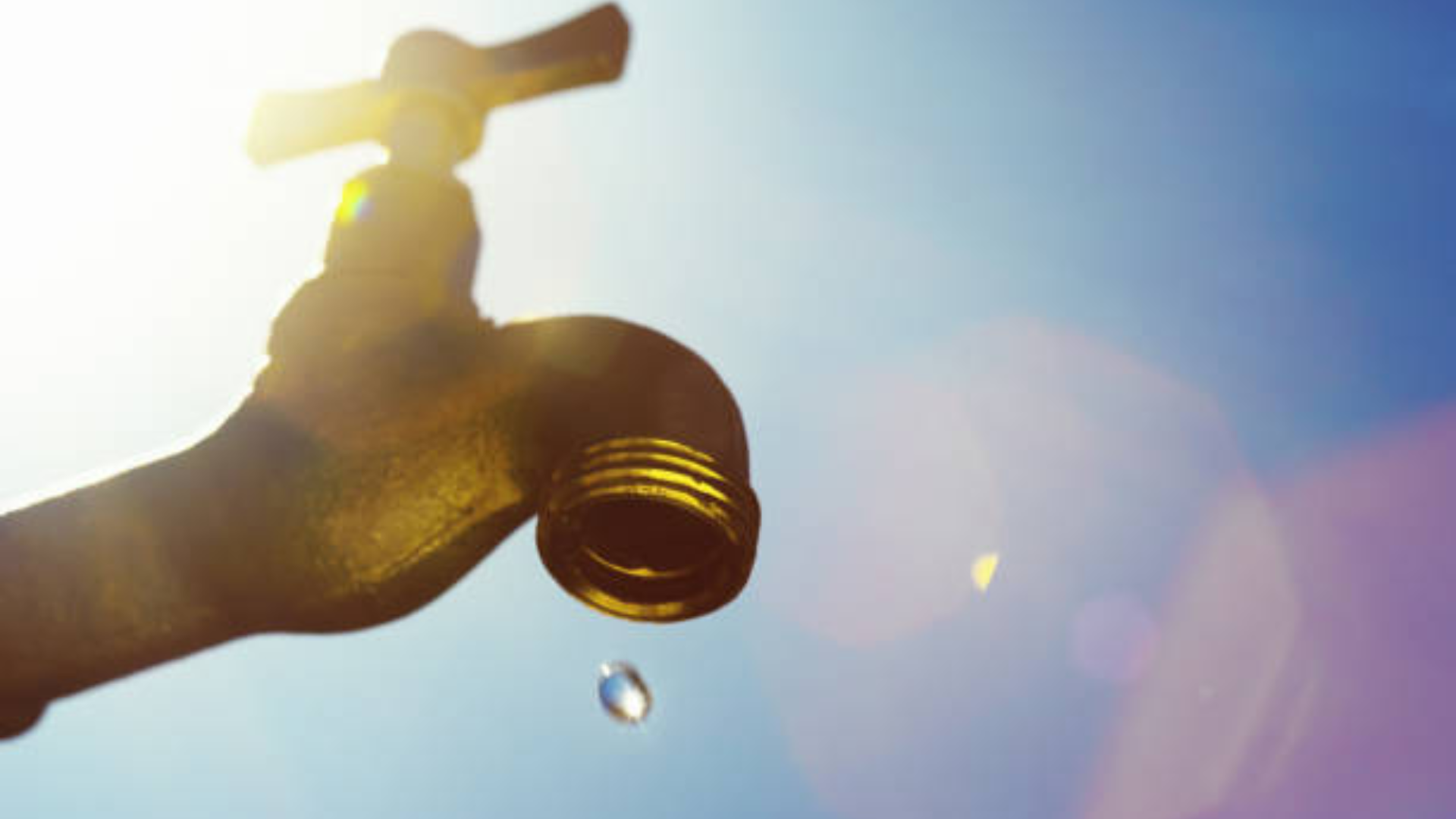 A low-angle silhouette of a metal outdoor water faucet with a single drop of water hanging from the spout.