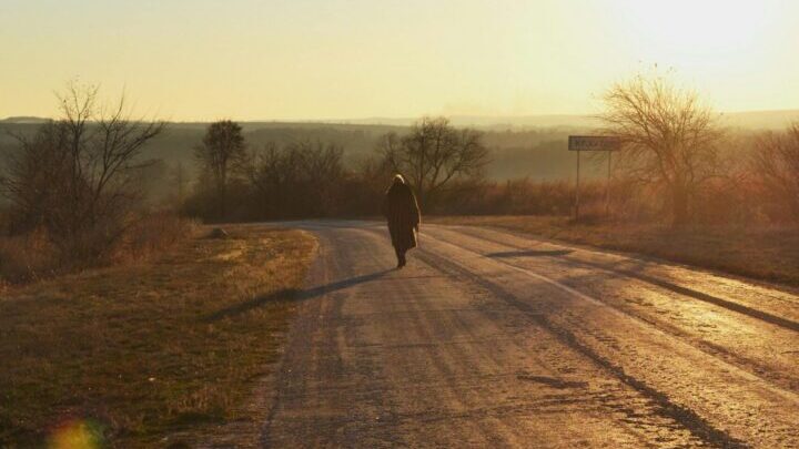 Person walking along a rural dirt road at sunrise with long shadows across the landscape.