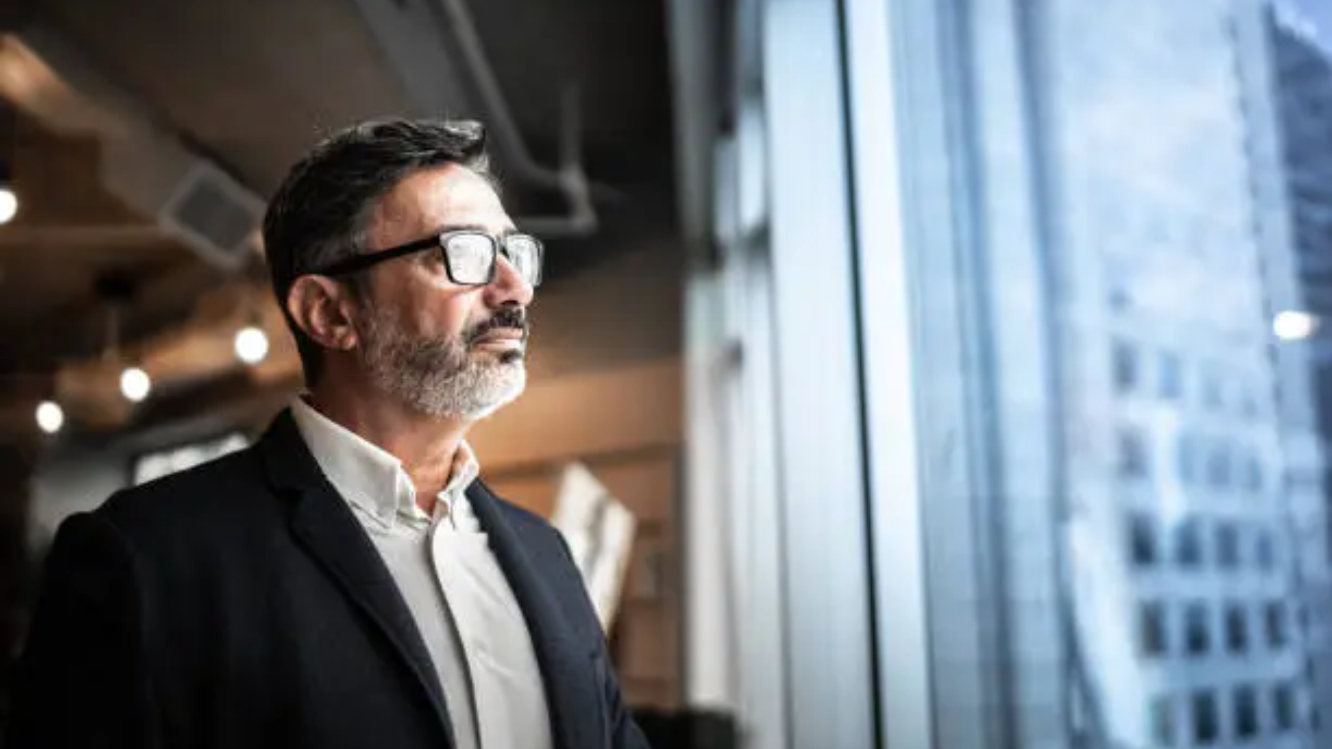 A professional medium shot of a middle-aged man with short dark hair and a salt-and-pepper beard wearing glasses and a dark blazer over a white shirt; he is looking thoughtfully out a large window toward a city skyline.