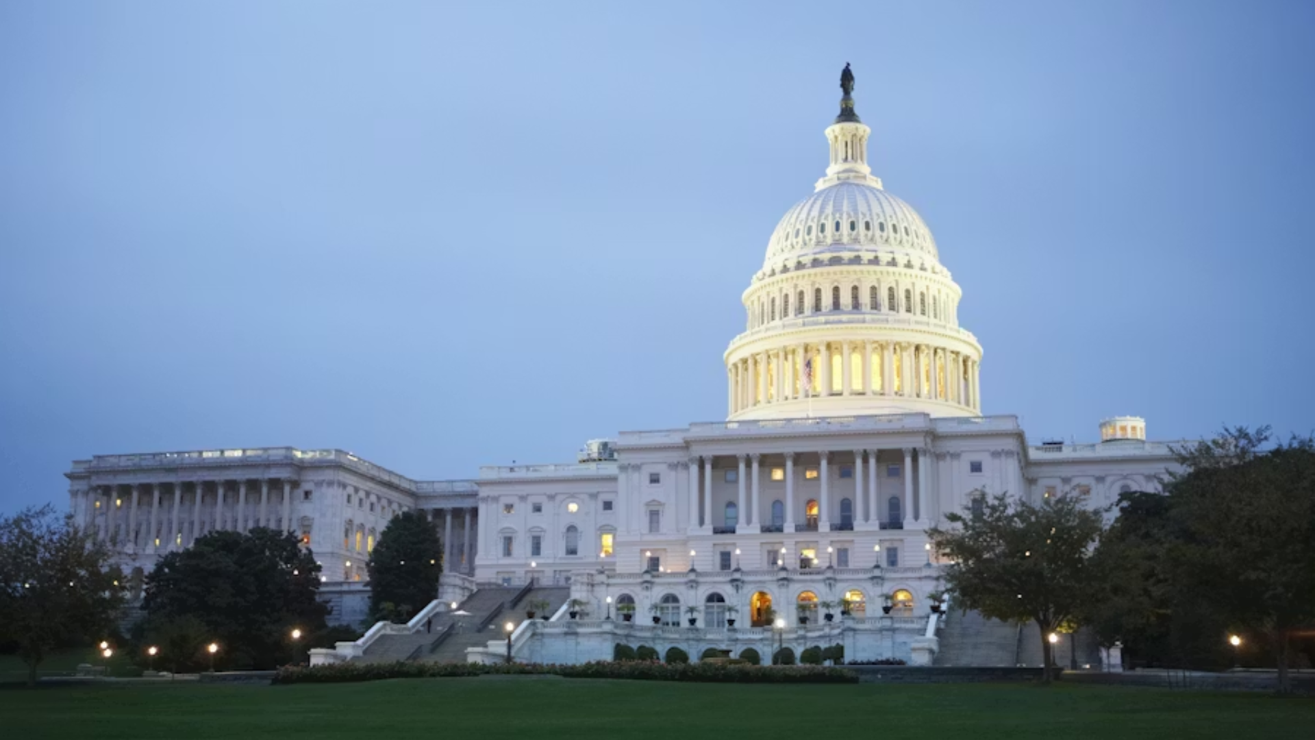 A wide daytime shot of the U.S. Capitol Building in Washington, D.C.; the iconic white dome is illuminated against a twilight sky, with the building's neoclassical architecture and the lush green lawn of the National Mall visible in the foreground.
