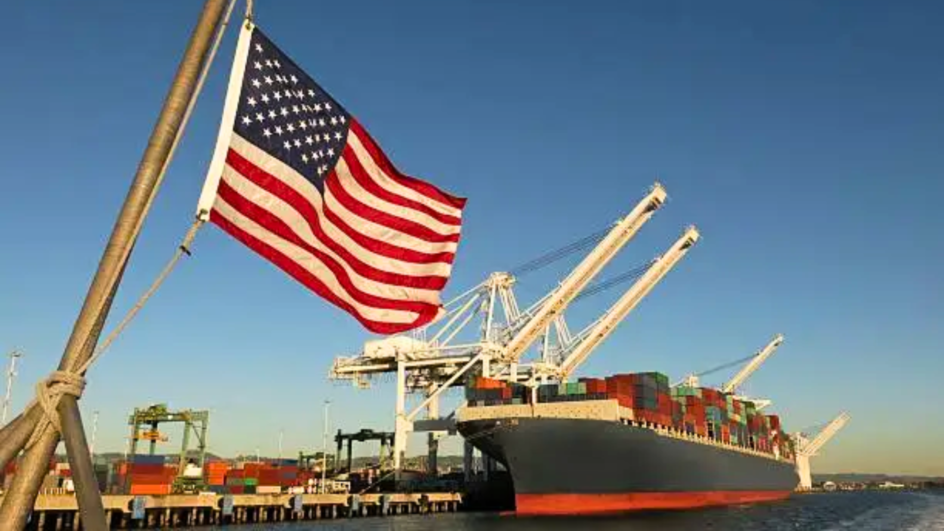 A low-angle shot of a large container ship docked at a harbor under a bright morning sun; the American flag is prominently displayed on a flagpole in the foreground.