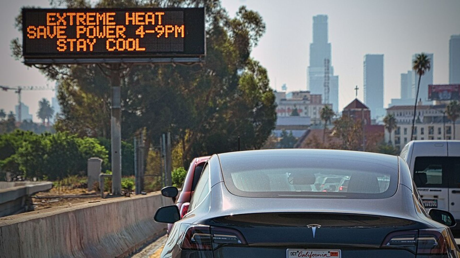 Digital road sign reading “Extreme Heat Save Power 4–9PM Stay Cool” above traffic with Los Angeles skyline in background.