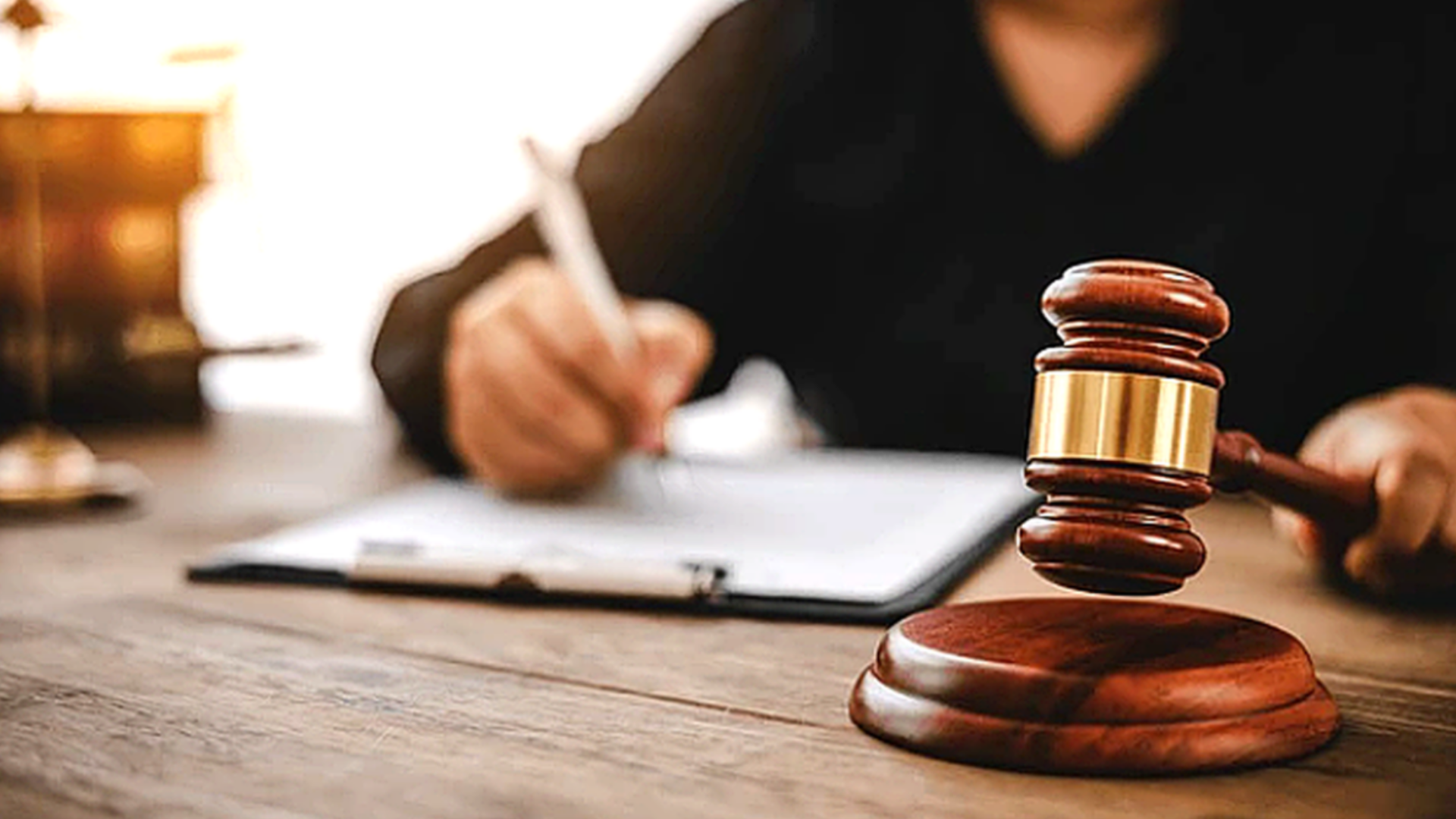 Judge’s gavel on a desk in foreground as a person signs documents in a courtroom setting.