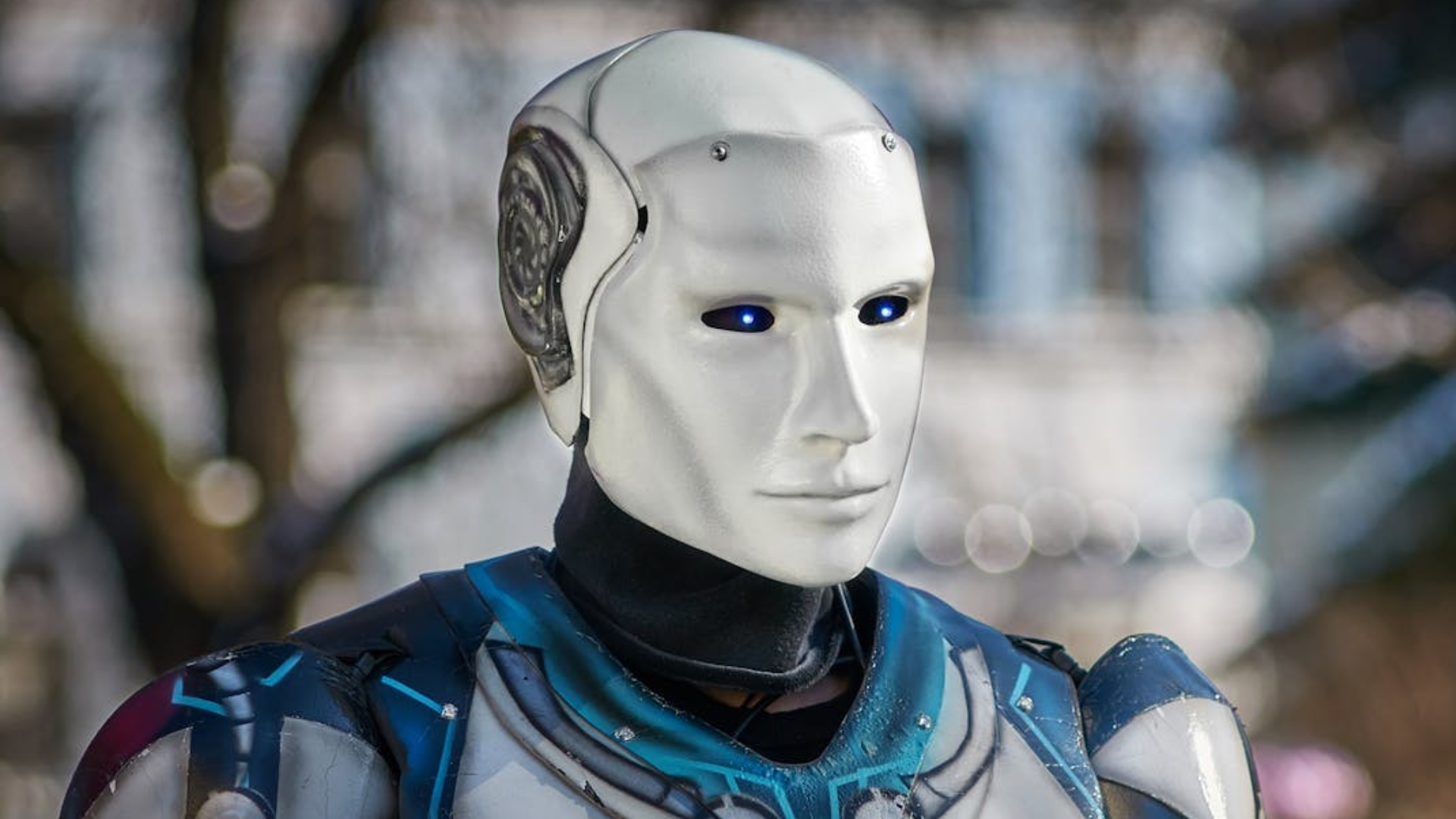 A humanoid robot with a smooth white face and glowing blue eyes stands outdoors.
